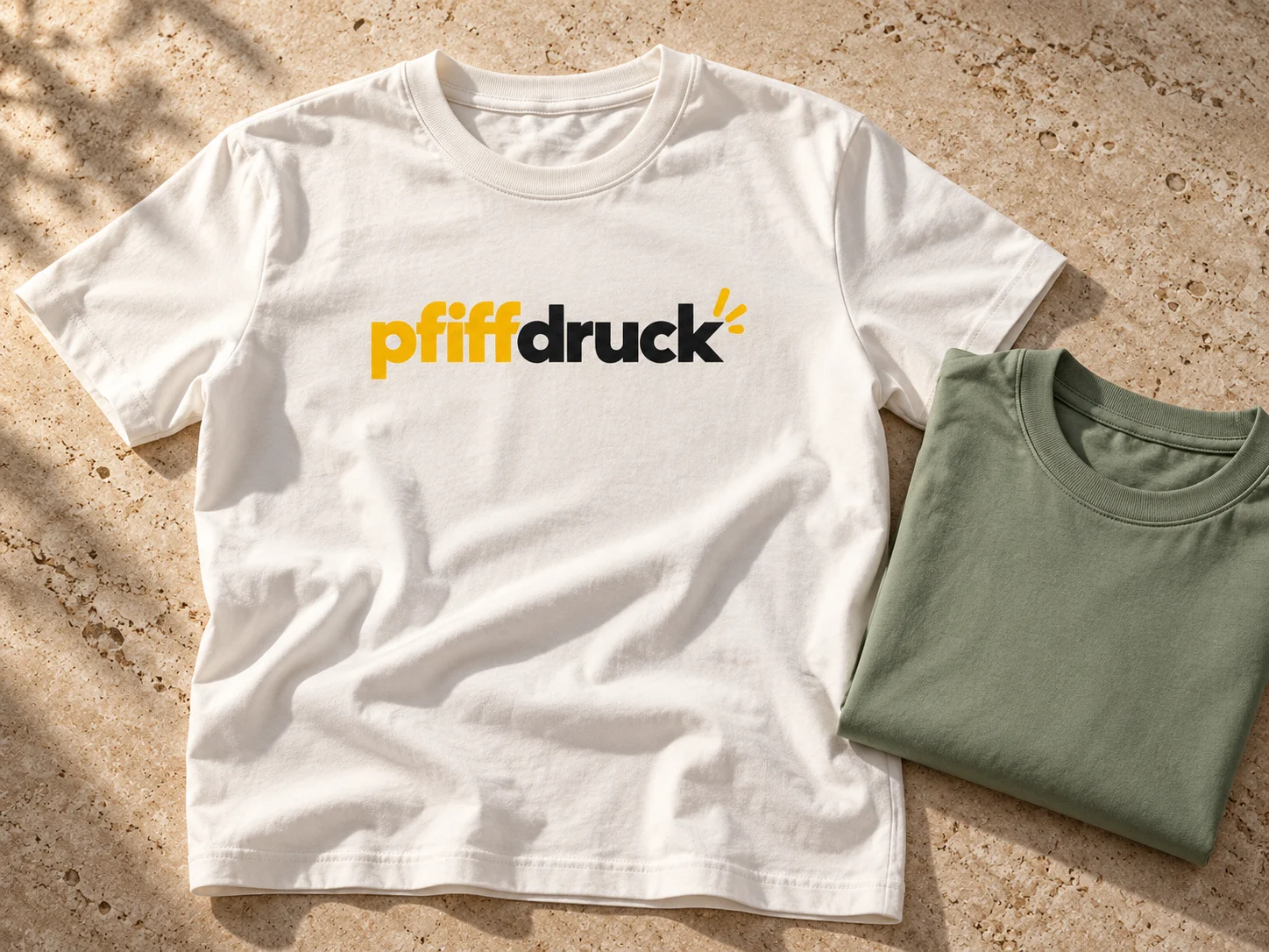Weißes T-Shirt mit pfiffdruck-Motiv neben einem grünen Shirt – Gestaltungsbeispiel
